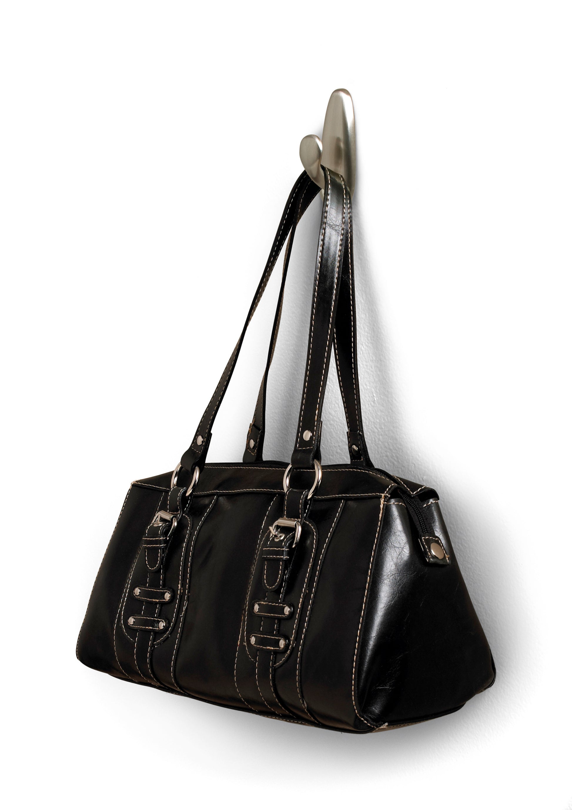 An einem Command™ Metallhaken "klassisch" der 3M Deutschland GmbH hängt eine schwarze Lederhandtasche mit Doppelgriffen und Zierschnallen. Die Tasche hat weiße Nähte und eine leicht glänzende Oberfläche.