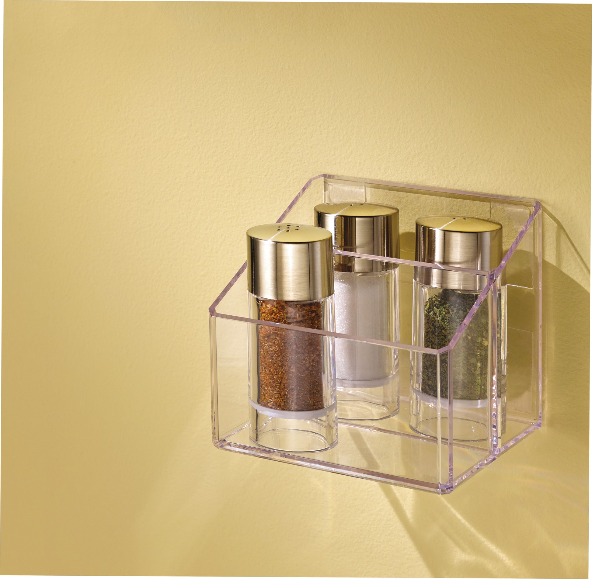 Drei gläserne Gewürzdosen mit goldenen Deckeln stehen in einem durchsichtigen Halter, der mit 3M Deutschland GmbH Scotch-FIX™ Transparentes doppelseitiges Montageband 4910C an einer hellgelben Wand befestigt ist. Die Gläser enthalten braune, weiße und grüne Gewürze.