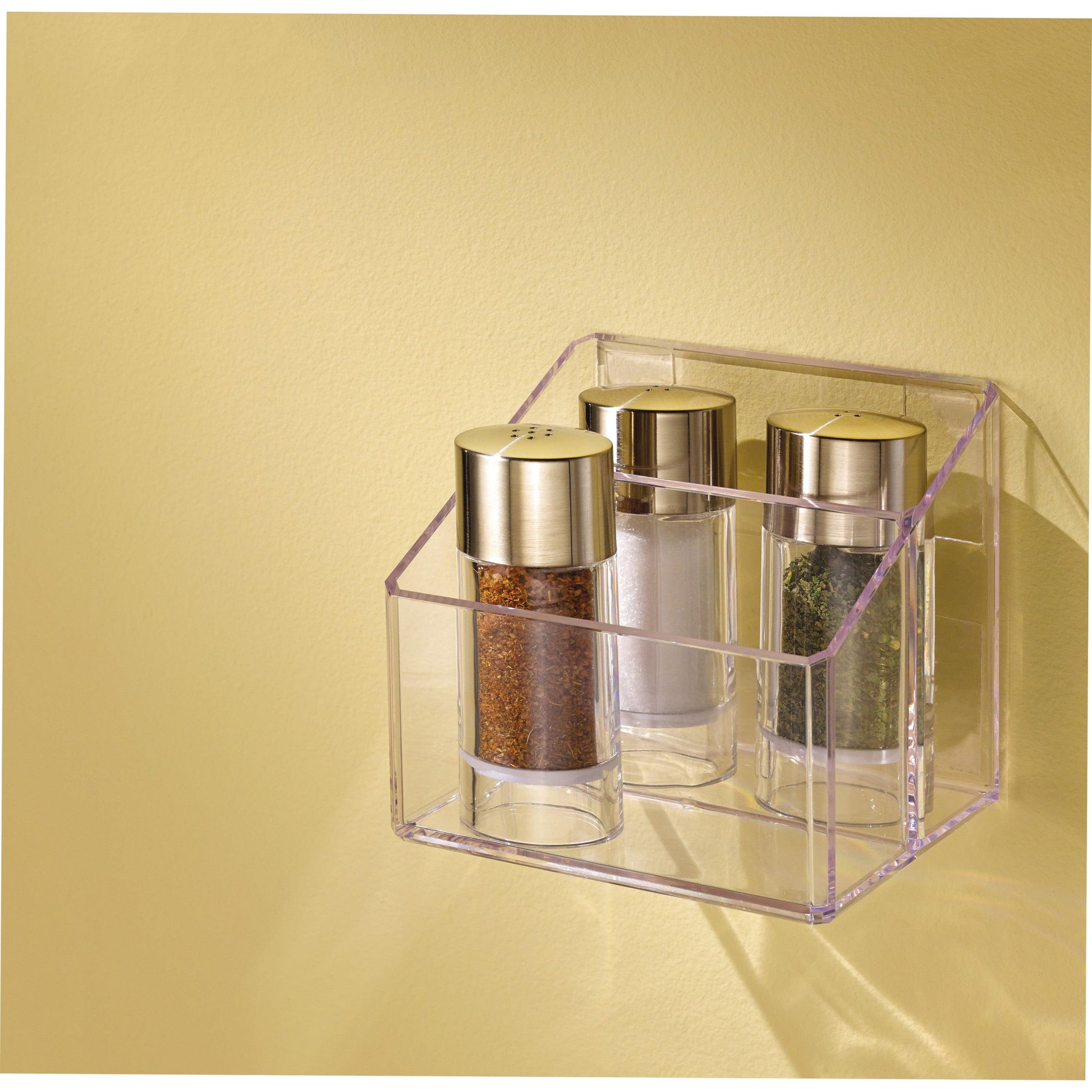 Drei gläserne Gewürzdosen mit goldenen Deckeln stehen in einem durchsichtigen Halter, der mit 3M Deutschland GmbH Scotch-FIX™ Transparentes doppelseitiges Montageband 4910C an einer hellgelben Wand befestigt ist. Die Gläser enthalten braune, weiße und grüne Gewürze.