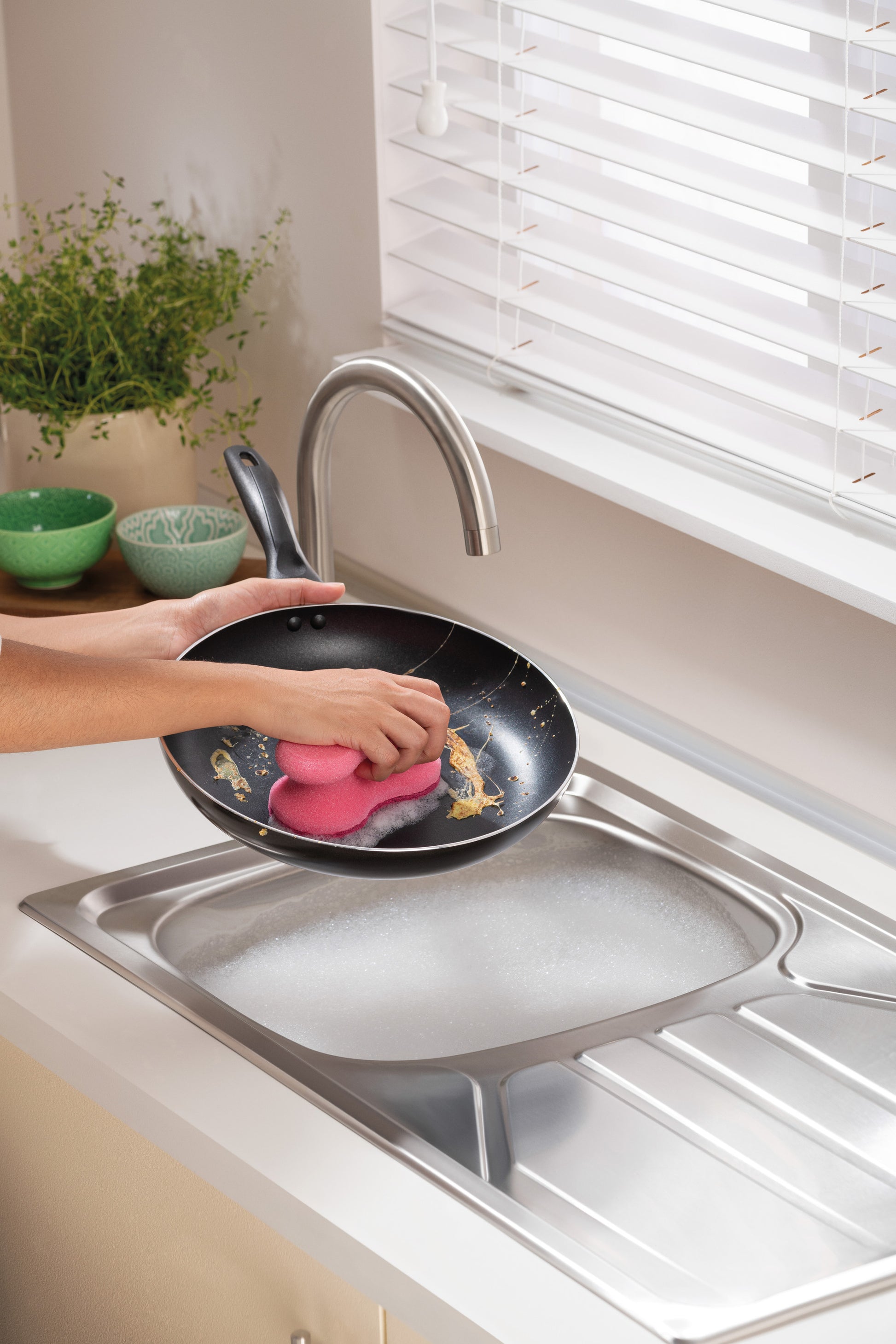 Eine Person reinigt eine schmutzige Bratpfanne mit einem rosa Scotch-Brite® Delicate Color Comfort nicht kratzender Reinigungsschwamm von 3M Deutschland GmbH unter einem Wasserhahn in einer seifigen Küchenspüle. Grüne Schalen und eine Topfpflanze stehen auf der Arbeitsplatte am Fenster.
