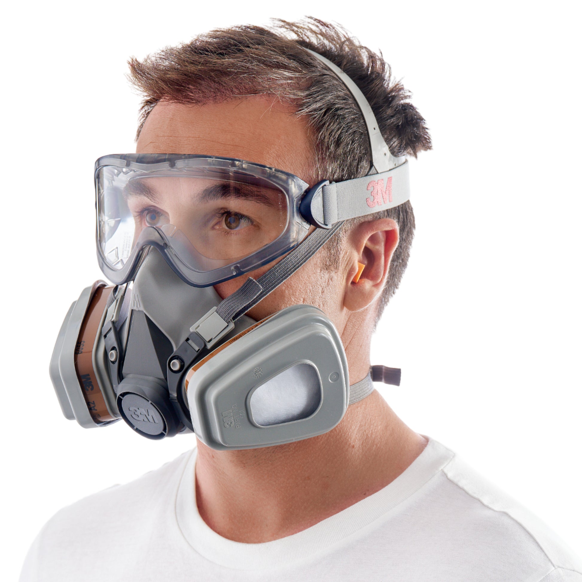 Ein Mann trägt die 3M™ Arbeitsschutz-Starter-Box für Profis 1000MCWE - ein Atemschutzgerät mit zwei Kartuschen, eine durchsichtige Schutzbrille und gelbe Gehörschutzstöpsel - von 3M Deutschland GmbH. Er hat kurzes braunes Haar und trägt ein weißes Hemd vor einem weißen Hintergrund.