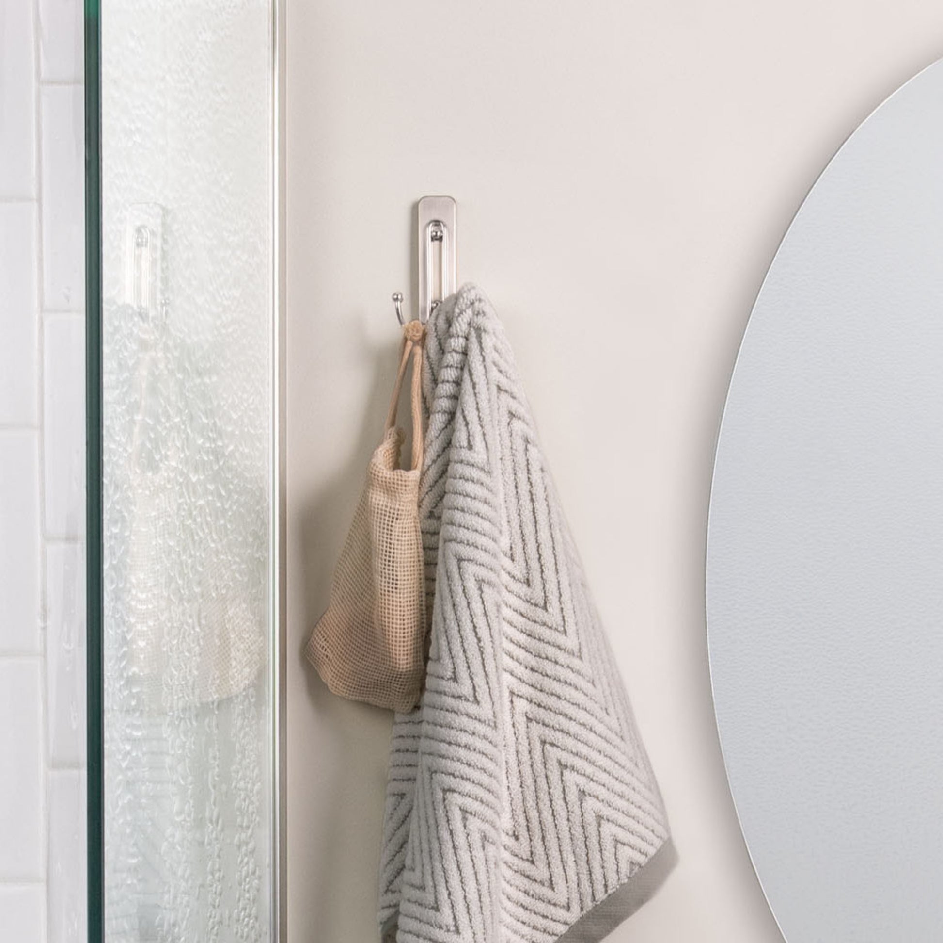 A Command™ Großer Badezimmer-Doppelhaken Nickel satiniert von 3M Deutschland GmbH hält ein graues, geometrisch gemustertes Handtuch und eine beige Mesh-Tasche neben einer Milchglasduschtür und einem runden Spiegel.