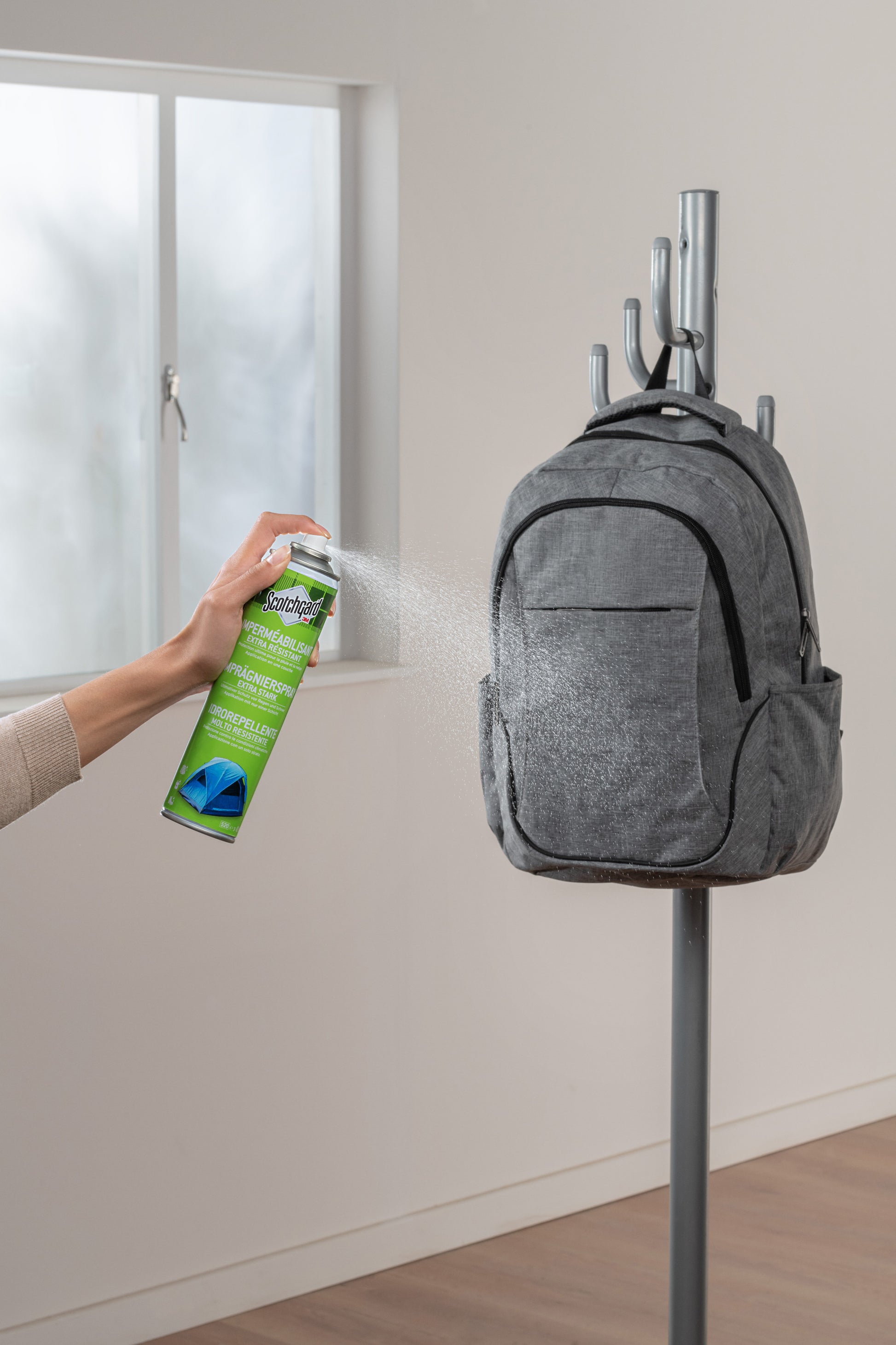 Eine Person sprüht Scotchgard™ Imprägnierspray extra stark (400ml, 1 Dose) von 3M Deutschland GmbH auf einen grauen Rucksack, der an einem Kleiderständer in der Nähe eines Fensters hängt.