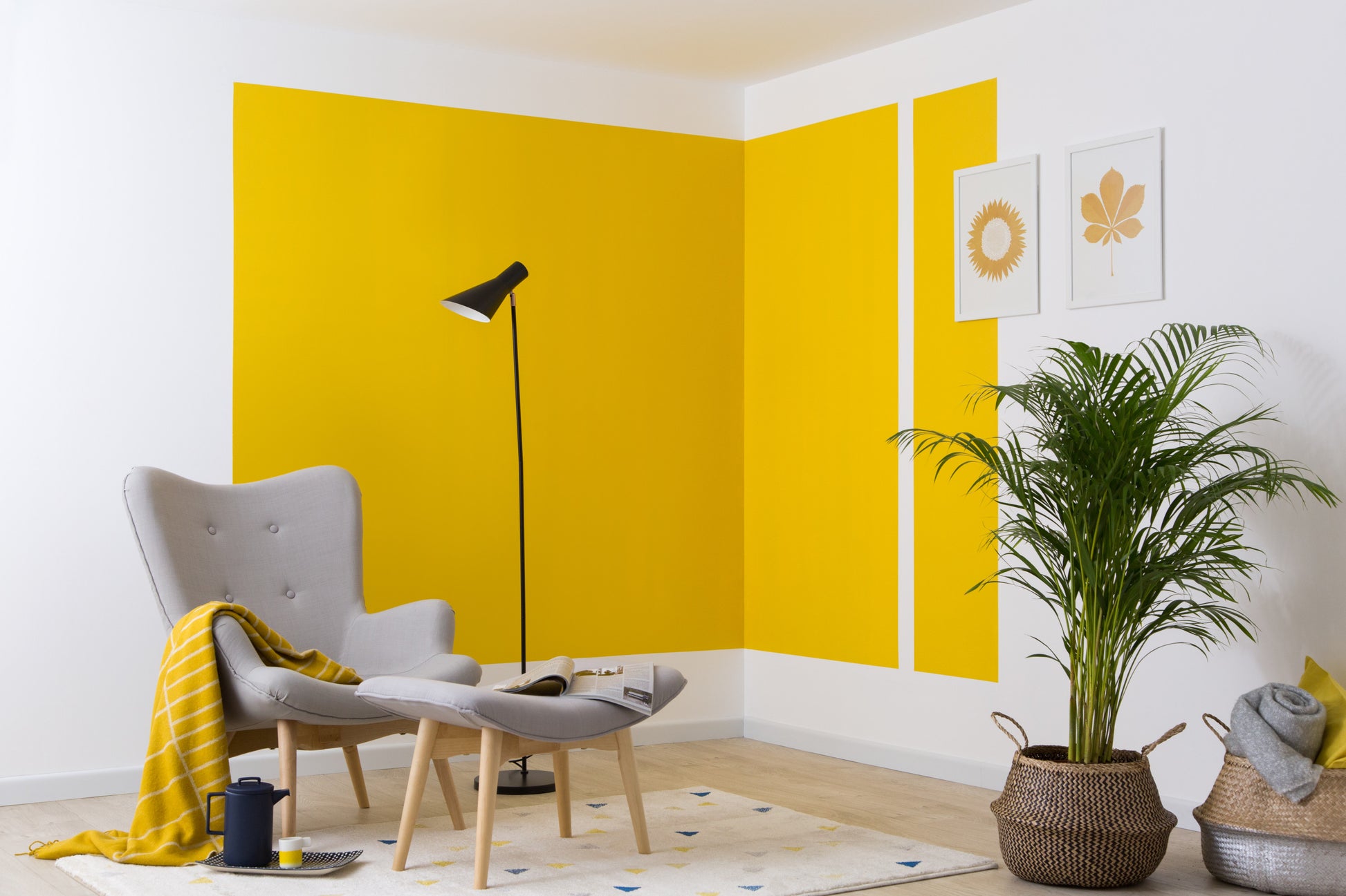 Ein modernes Wohnzimmer mit einem grauen Sessel, einer Ottomane, einer schwarzen Stehlampe, einer gelben Decke und einer Topfpflanze. Die kräftigen gelben geometrischen Formen an der Wand wurden mit 3M Deutschland GmbH ScotchBlue™ Malerabdeckband für scharfe Linien erstellt.