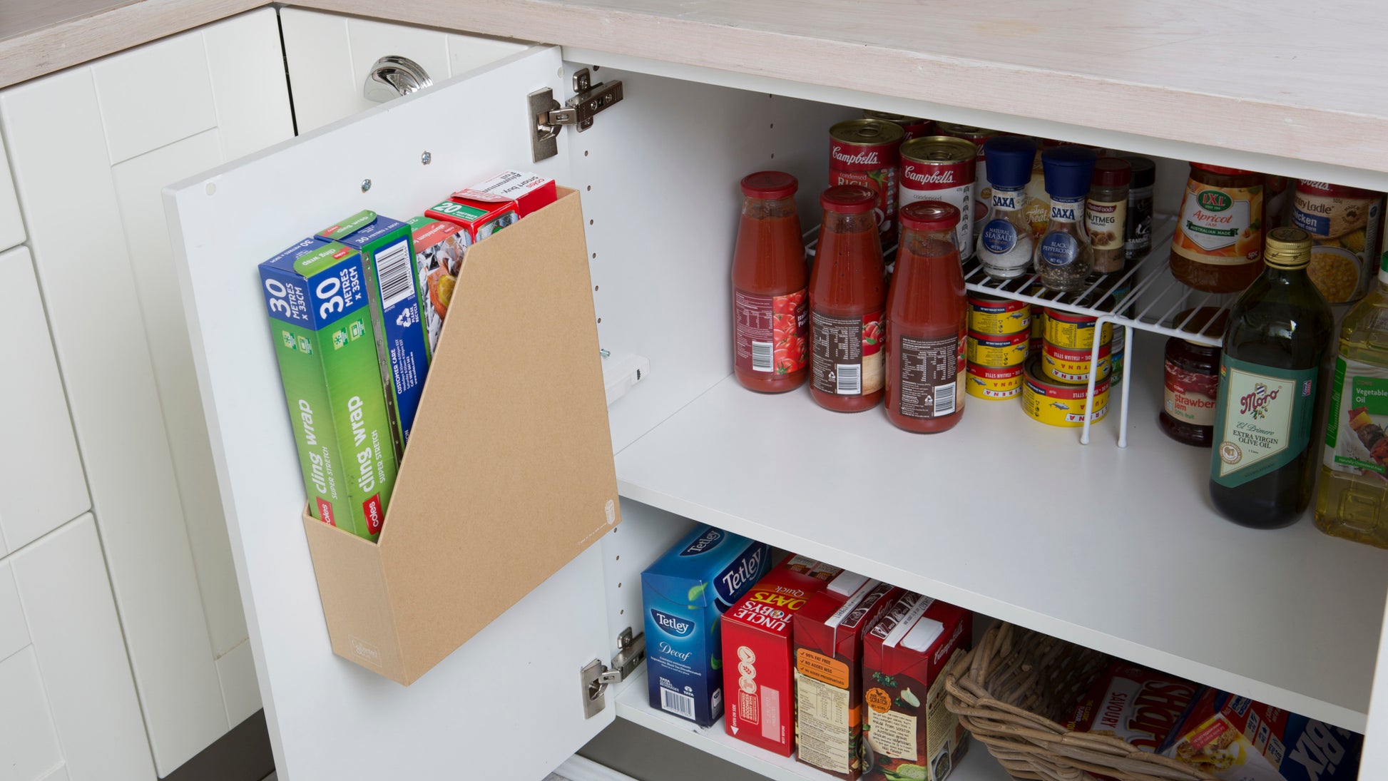 Ein Küchenschrank mit geöffneter Tür zeigt drei Müslischachteln in einer Halterung, die mit 3M Deutschland GmbH Scotch-FIX™ Doppelseitiges Innenmontageband an der Tür befestigt ist. Im Inneren befinden sich Gläser, Konserven, Flaschen und ein Korb auf dem unteren Regalboden.