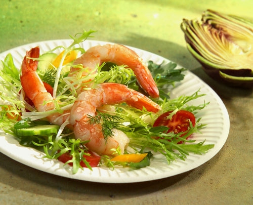 Ein PAPSTAR 100 Teller, Pappe "pure" rund, weiß extra stark der PAPSTAR GmbH hält einen Salat mit Blattgemüse, Garnelen, Gurken, gelber Paprika, Kirschtomaten und Dill. Im Hintergrund liegt ein aufgeschnittener Rotkohlkopf auf einer grünen Fläche.