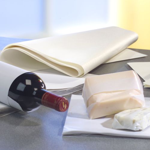 Eine Flasche Rotwein und Käseblöcke sind teilweise in PAPSTAR 10 kg Einschlagpapiere, Cellulose weiß der PAPSTAR GmbH eingewickelt, im Hintergrund ist auf der Tischplatte ein Stapel Wachspapierblätter zu sehen.