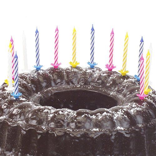 Ein mit Puderzucker bestreuter Schokoladen-Gugelhupf ist mit PAPSTAR 24 Geburtstagskerzen mit Halter (6 cm, farbig sortiert) verziert, die am Rand für eine festliche Optik sorgen. Marke: PAPSTAR GmbH.