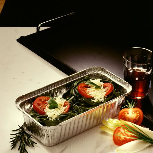 Eine Starpak 25 Schalen Aluminiumschale (PP-beschichtet mit Einsatzdeckel) ist mit ungekochten grünen Nudelnestern, Tomatenscheiben, Basilikum und geriebenem Käse auf einer weißen Fläche gefüllt. Daneben stehen ein Glas mit rotem Getränk, ganze Tomaten und Blattgemüse.