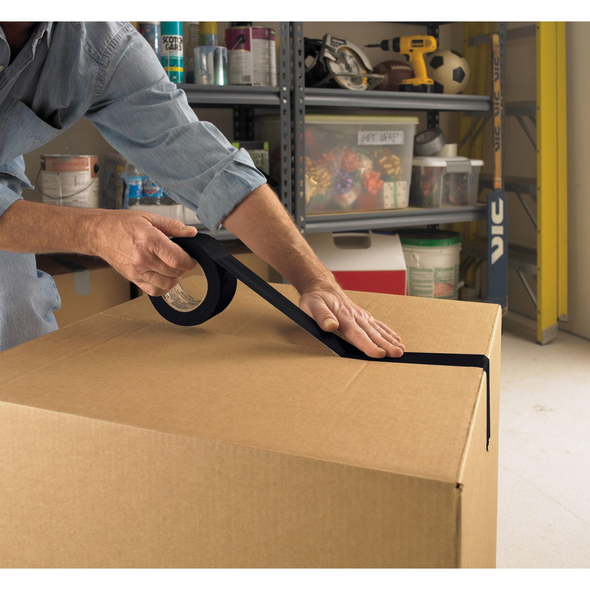 In einer Garage wird ein großer Karton mit Scotch® Extremium™ Universal Klebeband (3 m x 19 mm) der 3M Deutschland GmbH verschlossen, im Hintergrund sind Regale mit Haushaltsgegenständen zu sehen.