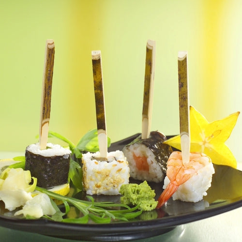 Drei Sushi-Stücke mit PAPSTAR 200 Fingerfood-Spieße 11 cm „Nature“ der PAPSTAR GmbH werden auf einem schwarzen Teller mit Wasabi, eingelegtem Ingwer, Blattgemüse und Sternfrucht angerichtet. Der unscharfe Hintergrund ist in Gelb- und Grüntönen gehalten.