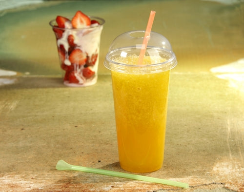 Ein Starpak 50 Trinkbecher "Hurricane", PET-Glasklar, mit gewölbtem Deckel und Strohhalm hält einen orangefarbenen Slush. Im Hintergrund steht ein weiterer Starpak Becher To-Go mit Erdbeeren und Sahne, daneben ein grüner Rührstab.