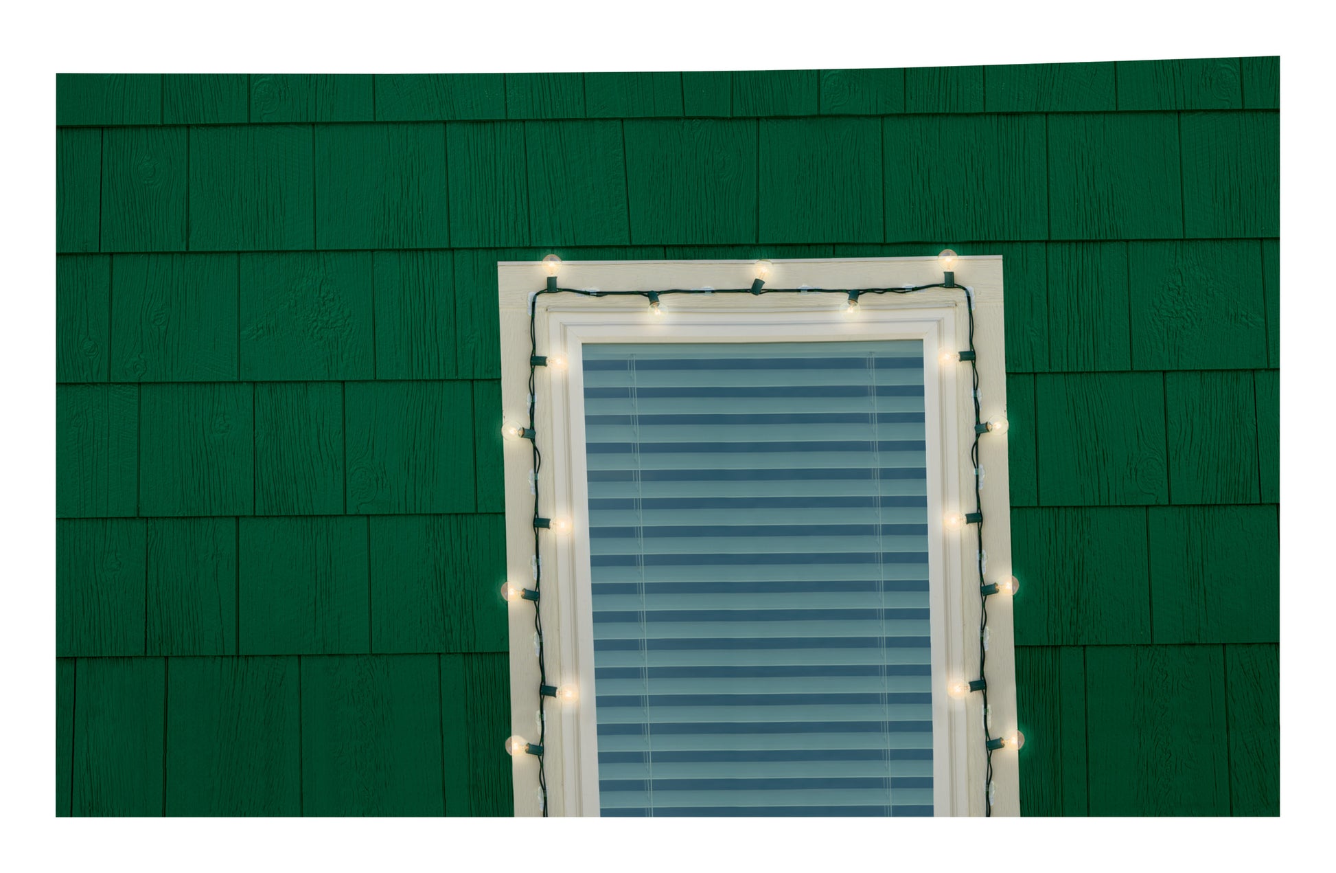 Eine grüne Wand zeigt ein Fenster mit geschlossenen Jalousien, eingerahmt von weißen Lichterketten, die mit 3M Deutschland GmbH Command™ Outdoor Lichter-Clips, 17017CLR-AWPU (8 Clips + 10 Streifen) aufgehängt wurden. Die Lichter sind eingeschaltet.