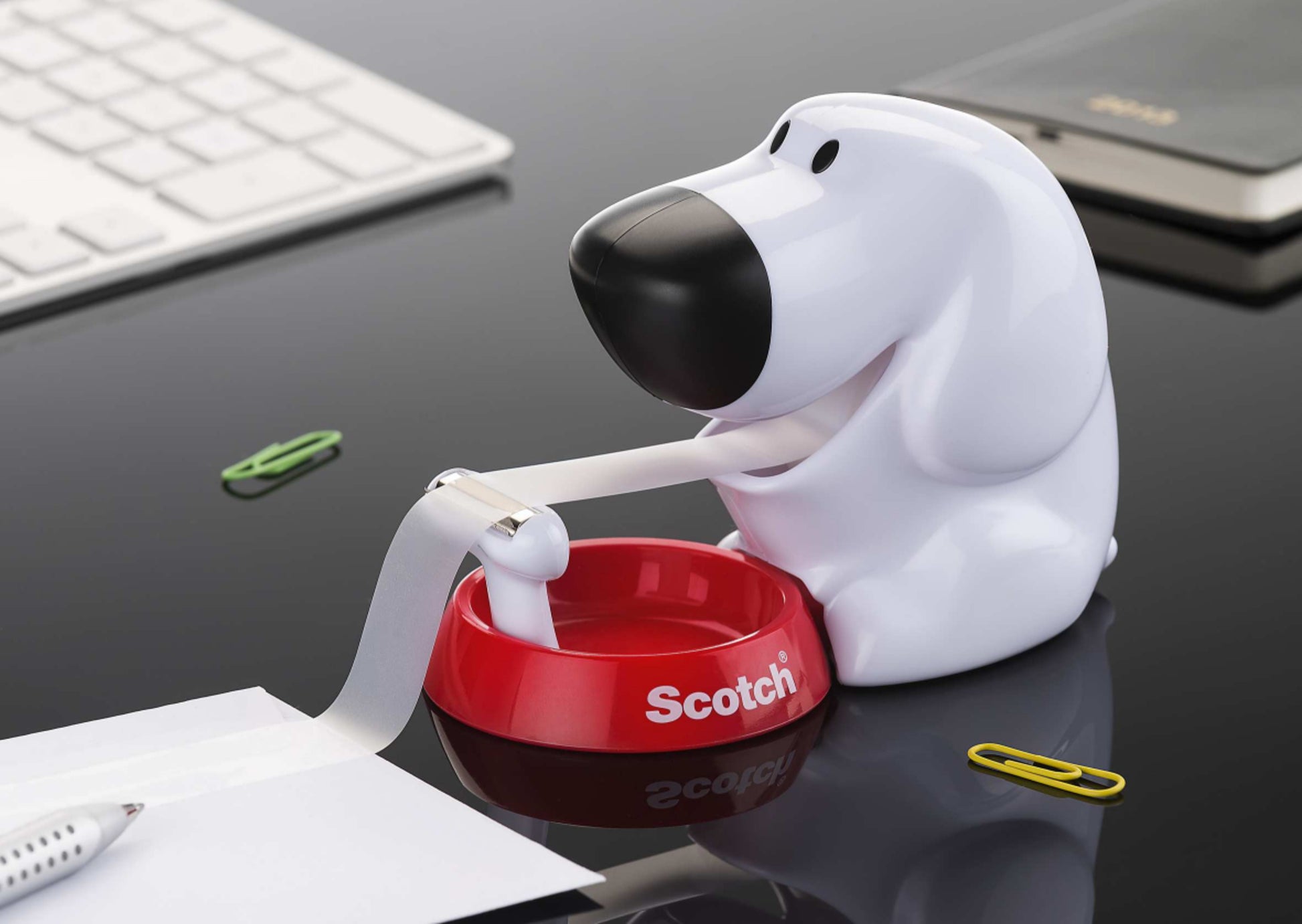 Der Scotch™ Tischabroller im Hunde Design C31 mit Scotch® Magic™ Klebeband (19 mm x 8,89 m, 1 Rolle) von 3M Deutschland GmbH steht auf einem schwarzen Schreibtisch neben Tastatur, Stift und Büroklammern.