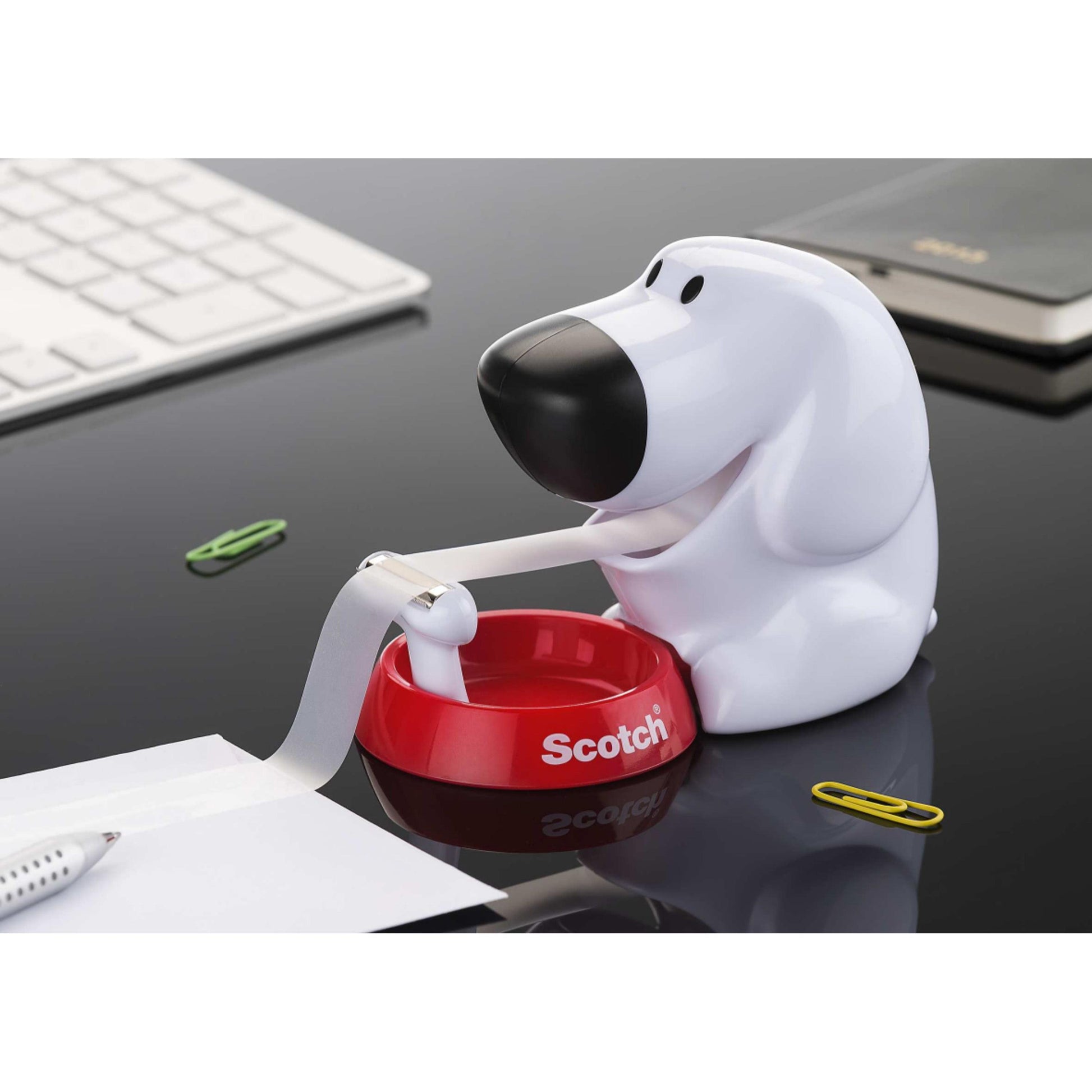 Der Scotch™ Tischabroller im Hunde Design C31 mit Scotch® Magic™ Klebeband (19 mm x 8,89 m, 1 Rolle) von 3M Deutschland GmbH steht auf einem schwarzen Schreibtisch neben Tastatur, Stift und Büroklammern.