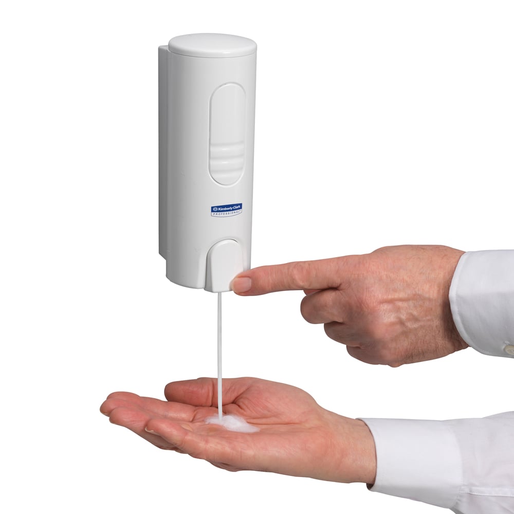 Eine Person gibt Kimberly-Clark Scott® ESSENTIAL™ Schaumseife (rosa, 200ml Kassette) aus einem weißen, an der Wand montierten Spender aus, indem sie mit einer Hand den Knopf der Seifenkassette drückt und den Schaum in der anderen Handfläche auffängt.