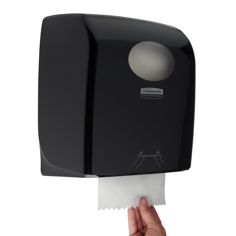 Eine Hand zieht ein Tuch aus dem Aquarius™ Rollenhandtuchspender - Schwarz von Kimberly-Clark GmbH. Der wandmontierte Spender hat ein transparentes Fenster, eine bebilderte Anleitung und ist ideal für den professionellen Einsatz. Größe: 38,20x30,90x24,00 cm.