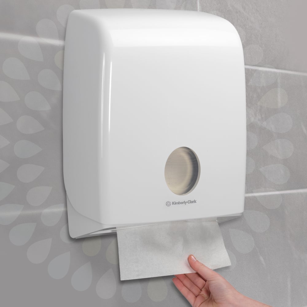 Eine Hand zieht ein Kleenex® Falthandtücher - Interfold aus einem weißen Wandspender der Kimberly-Clark GmbH in einem gefliesten Badezimmer. Der Spender hat ein rundes Fenster, das den Blick auf die Papierhandtücher im Inneren freigibt (15 Clips x 186 Blatt).
