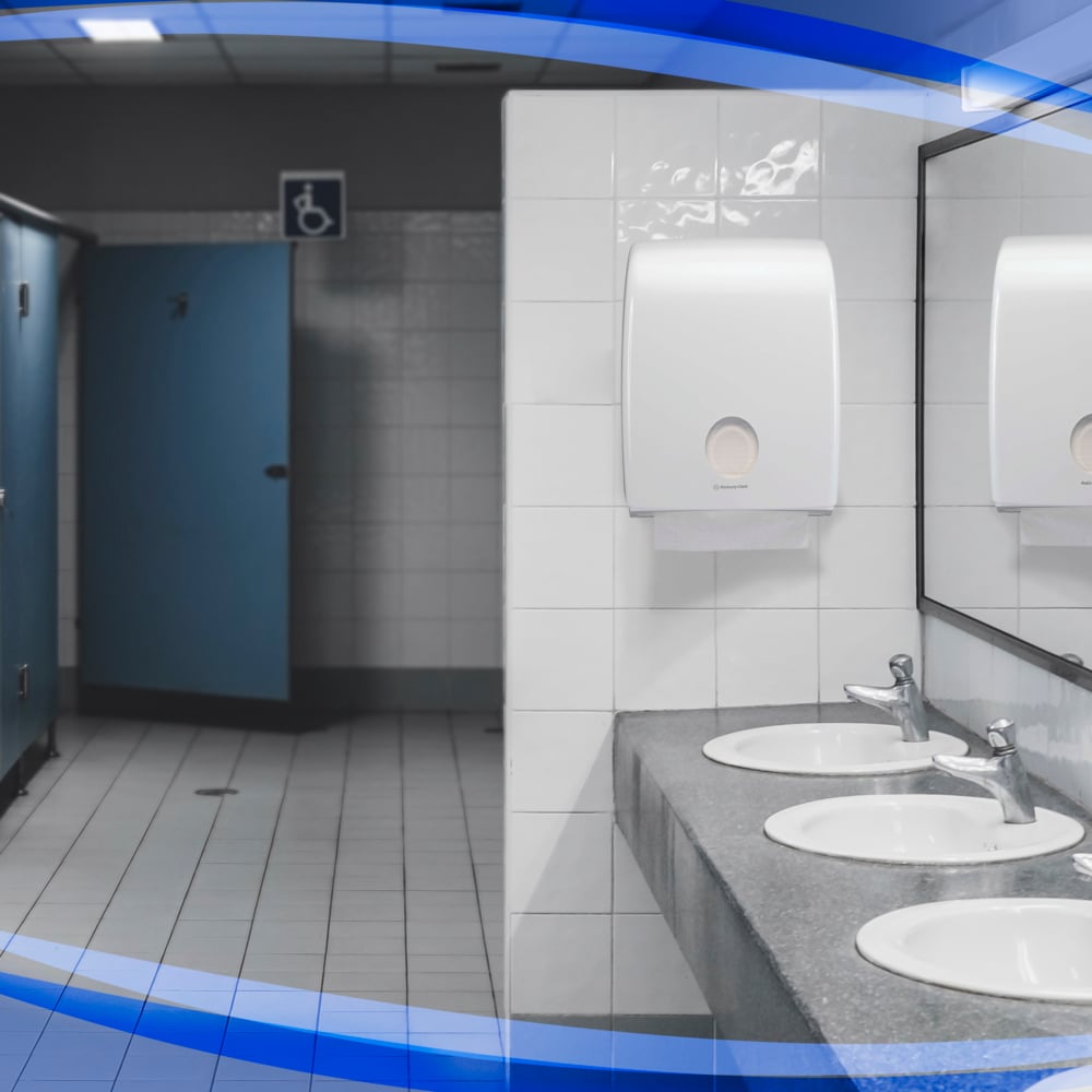 Eine öffentliche Toilette mit drei Waschbecken, Kimberly-Clark Aquarius™ Spender für Falthandtücher, hygienischen Seifenspendern, Spiegeln auf der rechten Seite und blauen Kabinen auf der linken Seite - eine offene Kabine ist für Rollstuhlfahrer zugänglich.