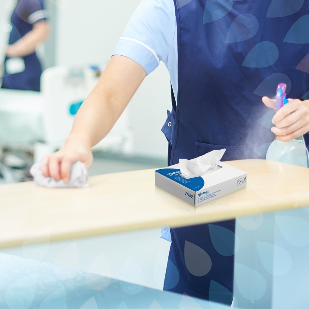 Eine Person in einer blauen Uniform reinigt einen hellen Tisch mit einem Tuch und einer Sprühflasche für die hygienische Reinigung. Eine Schachtel Kleenex® Wischtücher - Interfold / Weiß von Kimberly-Clark GmbH steht auf dem Tisch, im Hintergrund sind medizinische Geräte zu sehen.