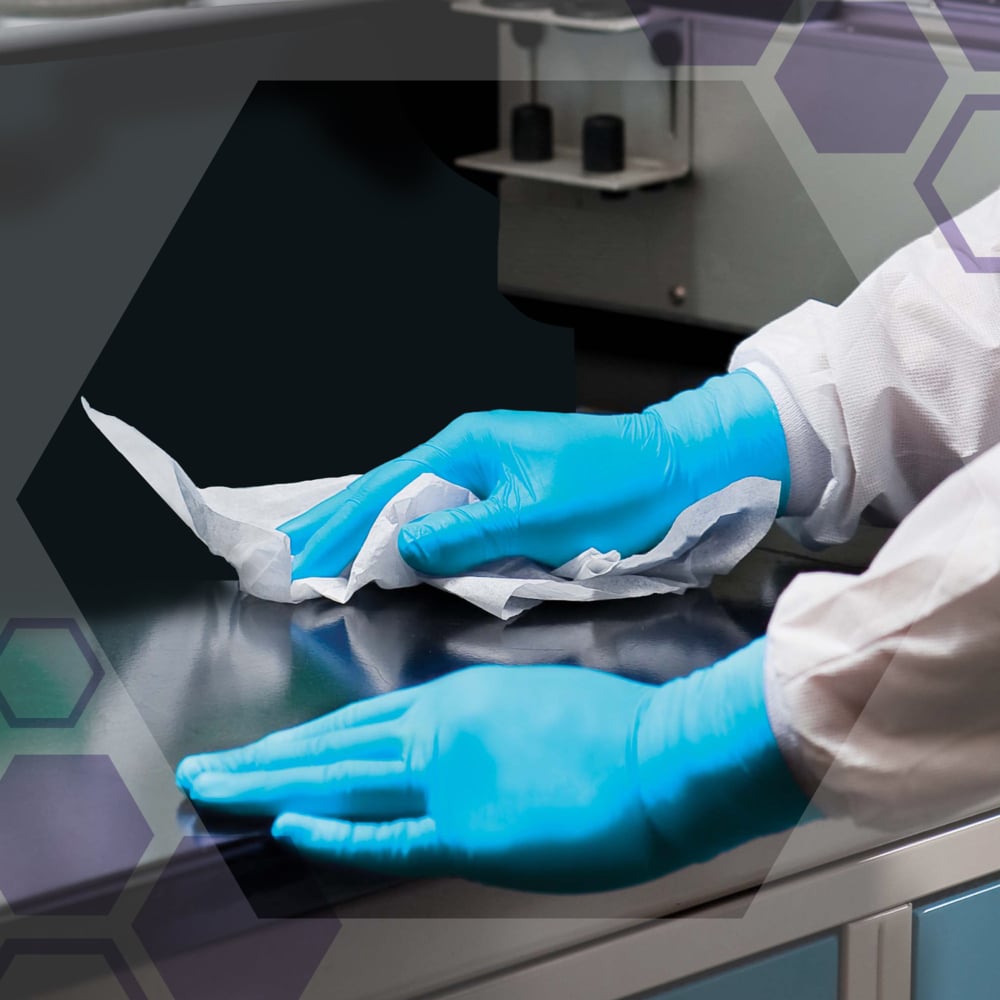 Eine Person mit blauen Handschuhen und einem weißen Laborkittel wischt eine dunkle Labortischplatte mit Kimtech® Saugfähige Pflegetücher, Weiß (20 Beutel x 50 Blatt, Karton) von Kimberly-Clark GmbH ab. Im Hintergrund sind Laborgeräte zu sehen.
