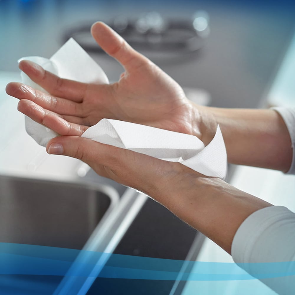 Eine Person trocknet ihre Hände über einem Spülbecken mit Scott® Handtüchern - MultiFold / Weiß (16 Clips x 250 Blatt | Karton) von Kimberly-Clark GmbH, mit unscharfen Arbeitsplatten und Küchenobjekten im Hintergrund.