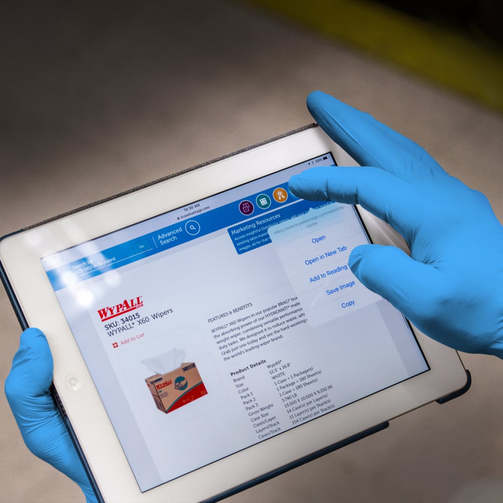 Eine Person, die KleenGuard® G10 2PRO Blaue Nitril-Handschuhe (23cm, beidhändig) von Kimberly-Clark GmbH trägt, verwendet ein Tablet, auf dem die Produktseite der WypAll X60 Wischtücher (SKU 34955) mit Details, Optionen und einem Bild der Verpackung angezeigt wird.