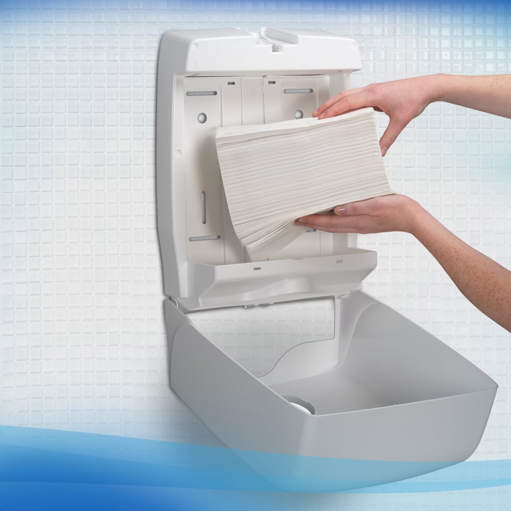 Eine Person lädt Scott® Control™ Herunterspülbare Falthandtücher von Kimberly-Clark GmbH in einen offenen Wandspender in einem weiß gefliesten Waschraum und sorgt so für hygienische Handtrocknung.