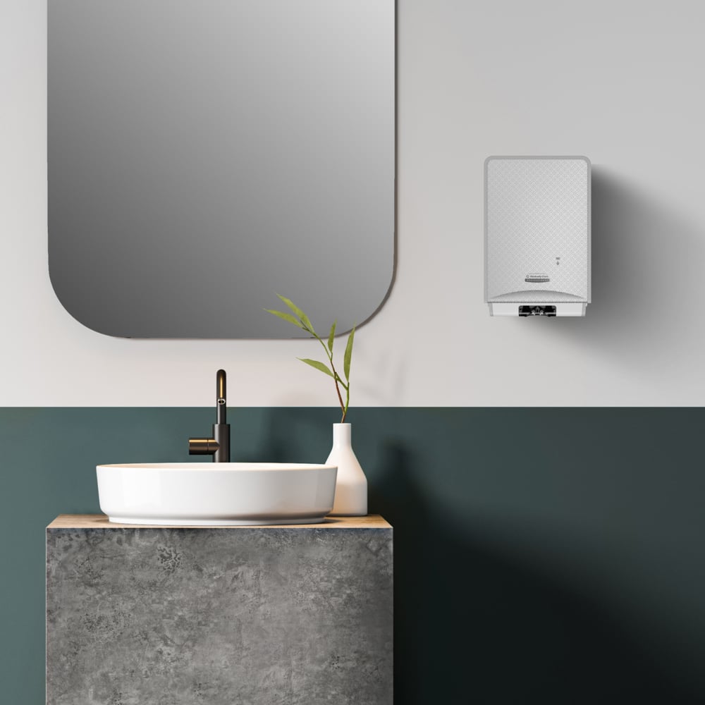 Ein modernes Badezimmer mit einem runden weißen Waschbecken auf einer Betontheke, einem schwarzen Wasserhahn, einer weißen Vase mit grünen Stielen, einem großen rechteckigen Spiegel und einer an der Wand montierten Kimberly-Clark Kleenex® Botanics™ Luxury Foam Hand Wash 1.2L-Kassette.