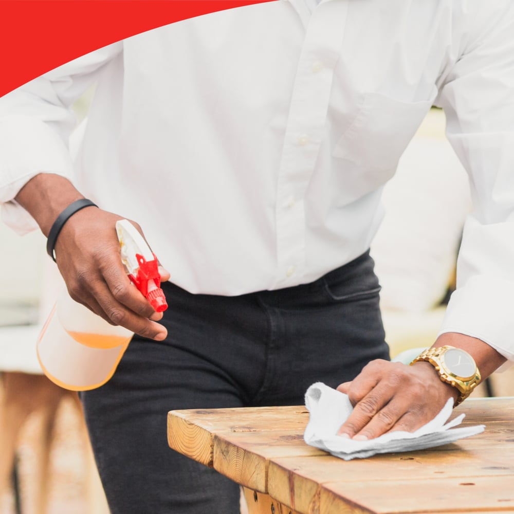 Eine Person in weißem Hemd und schwarzer Hose benutzt eine Sprühflasche und eine WypAll® L40 PowerClean Großrolle (Kimberly-Clark GmbH), um einen Holztisch zu reinigen. Die Person trägt eine Uhr; der obere Rand des Bildes zeigt eine rote geschwungene Umrandung.
