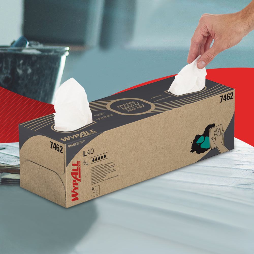 Eine Hand zieht ein WypAll® L40 PowerClean Wischtuch aus einer rechteckigen weißen Zupfbox der Kimberly-Clark GmbH, die auf einer Fläche steht, auf der ein weiteres Blatt zu sehen ist; die Box ist mit schwarzem und rotem Text und Abbildungen versehen.