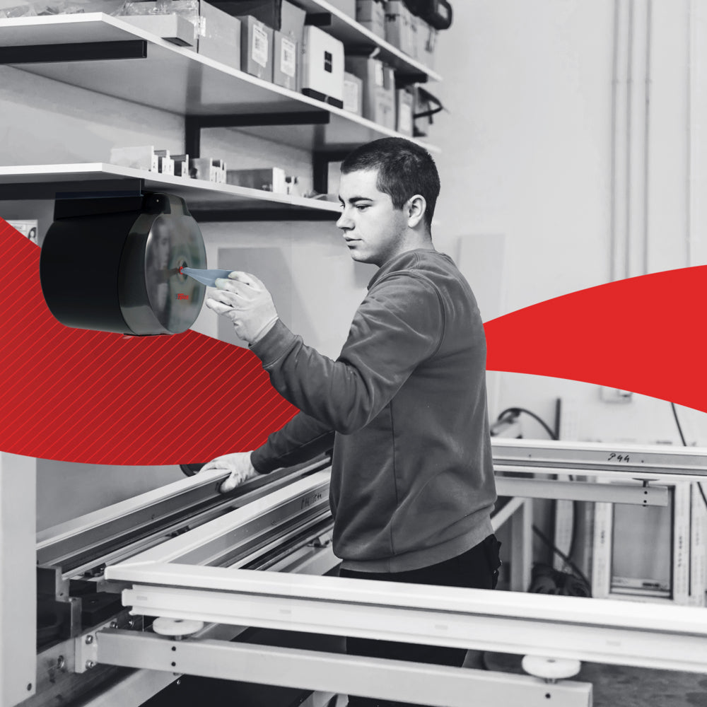 In einer Werkstatt richtet eine Person ein Maschinenteil neben einer Werkbank ein, die mit dem WypAll® L10 Papierwischtuch Centrefeed for Reach+™ (38,00 x 18,30 cm) der Kimberly-Clark GmbH für die schnelle Reinigung des Einzelhandels ausgestattet ist; im Hintergrund sind Regale und eine rote Grafik zu sehen.