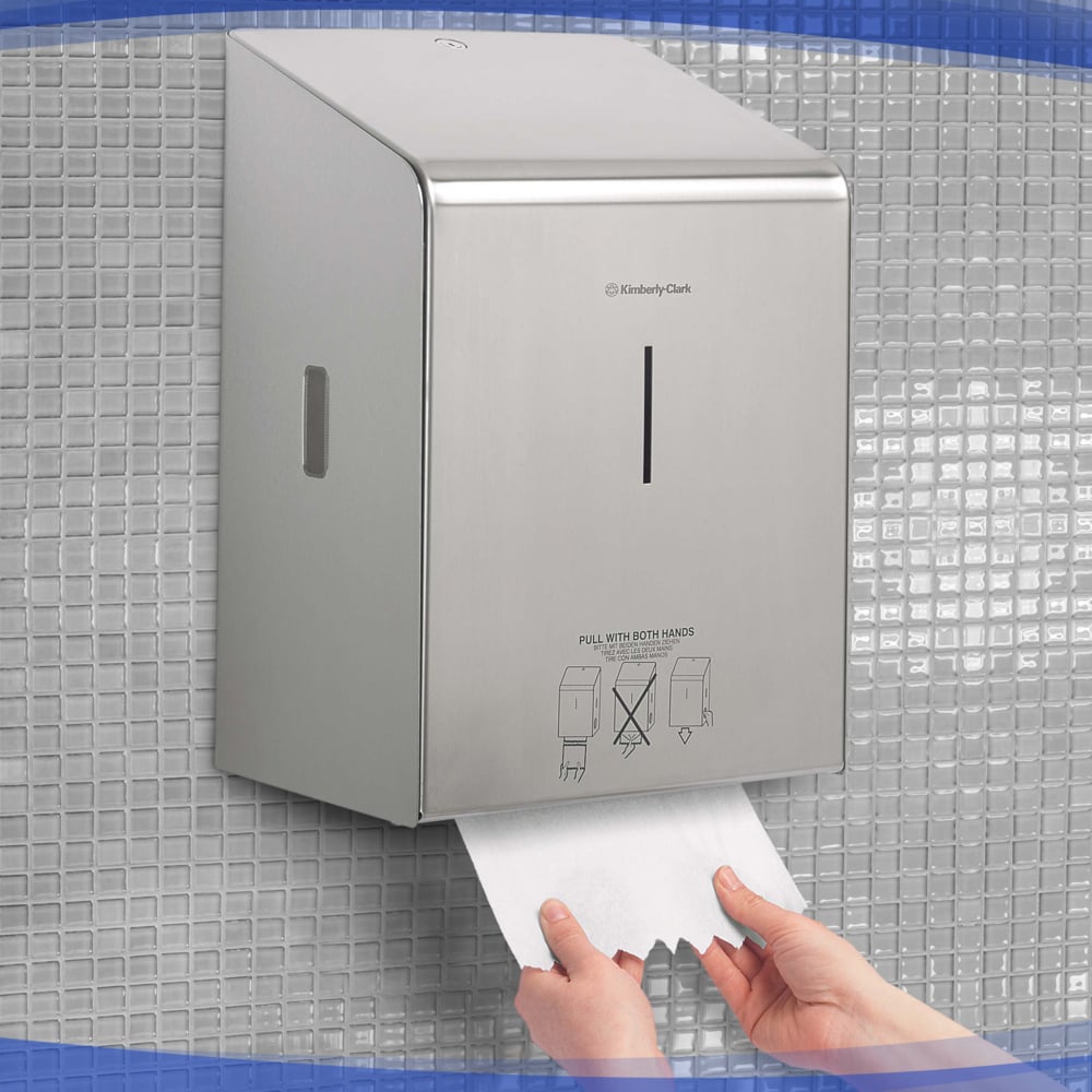 Eine Person zieht Papierhandtücher aus einem Kimberly-Clark Professional™ Rollenhandtuchspender - Edelstahl von Kimberly-Clark GmbH, der inmitten kleiner grauer quadratischer Fliesen an der Wand angebracht ist.