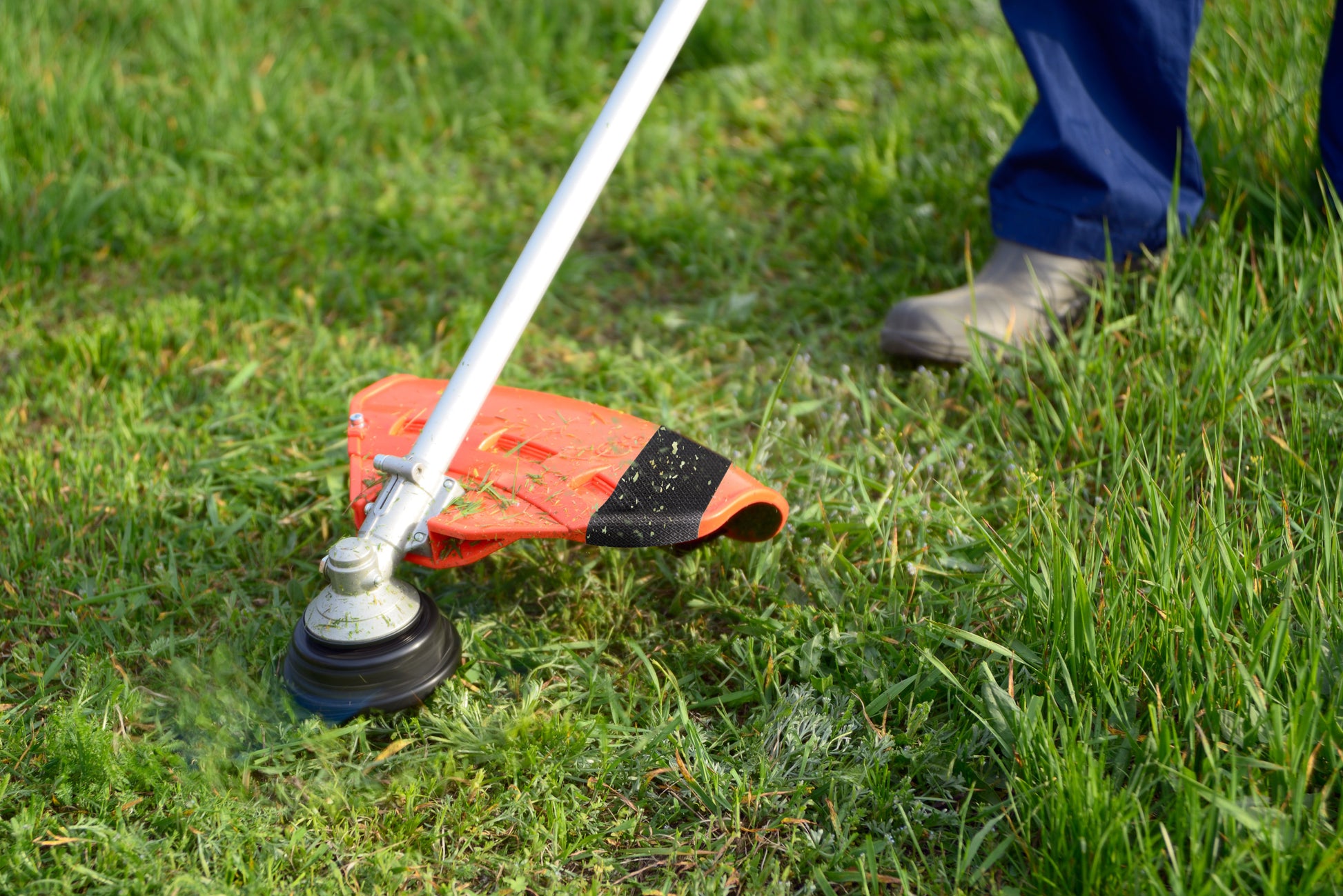 Mit festen Stiefeln trimmt eine Person das grüne Gras im Garten, während eine Rolle Scotch® Extremium™ Ultra Hochleistungs-Klebeband DT17 (10 m x 24 mm) von 3M in der Nähe liegt, bereit für anspruchsvolle Reparaturen.