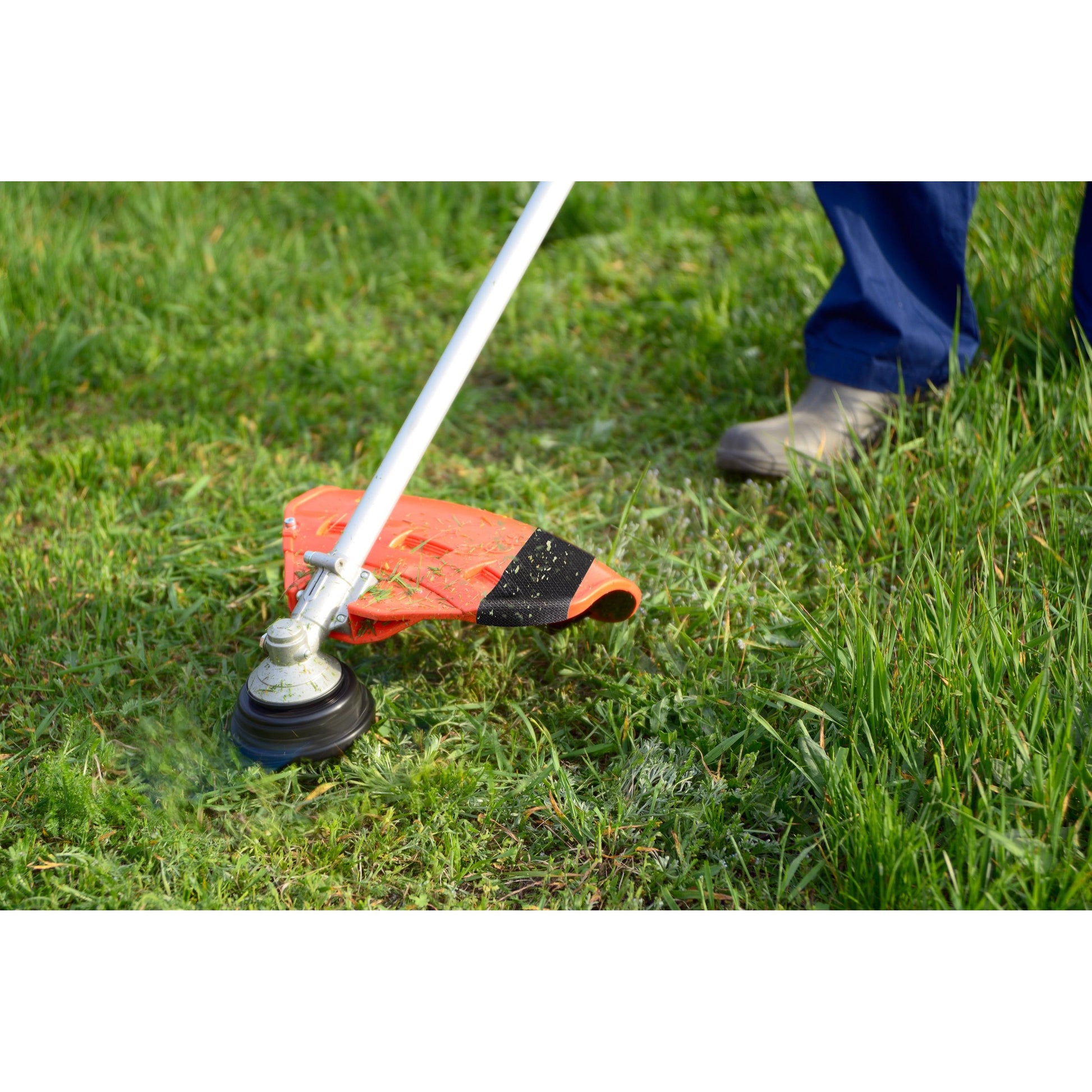 Mit festen Stiefeln trimmt eine Person das grüne Gras im Garten, während eine Rolle Scotch® Extremium™ Ultra Hochleistungs-Klebeband DT17 (10 m x 24 mm) von 3M in der Nähe liegt, bereit für anspruchsvolle Reparaturen.