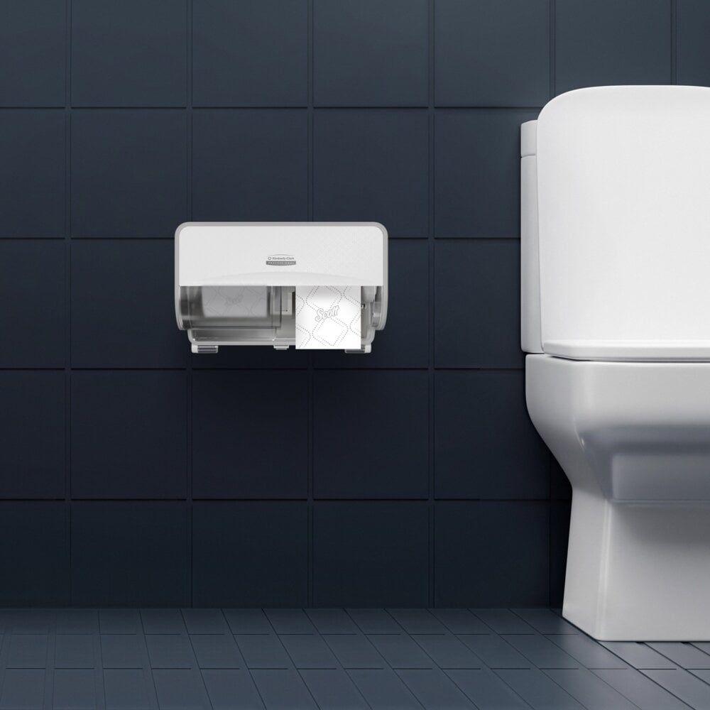 Ein wandmontierter Spender enthält zwei Rollen Scott® Essential™ Toilettenpapier 8517 2-lagig von Kimberly-Clark GmbH und ist an einer dunkel gefliesten Wand neben einer weißen Toilette mit geschlossenem Deckel angebracht; der Boden ist passend dazu mit dunklen Fliesen versehen.