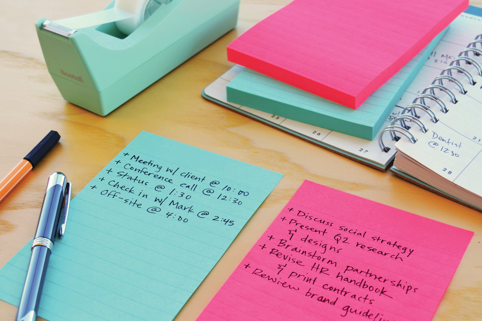 Zwei Stapel 3M Deutschland GmbH Post-it® Super Sticky Notes im Großformat (101 mm x 101 mm, 70 Blatt/Block, 100% PEFC) liegen auf einem Schreibtisch neben einem Stift, einem Klebebandabroller und einem aufgeschlagenen Planer, auf dem handschriftlich geplante Aufgaben und Termine eingetragen sind.