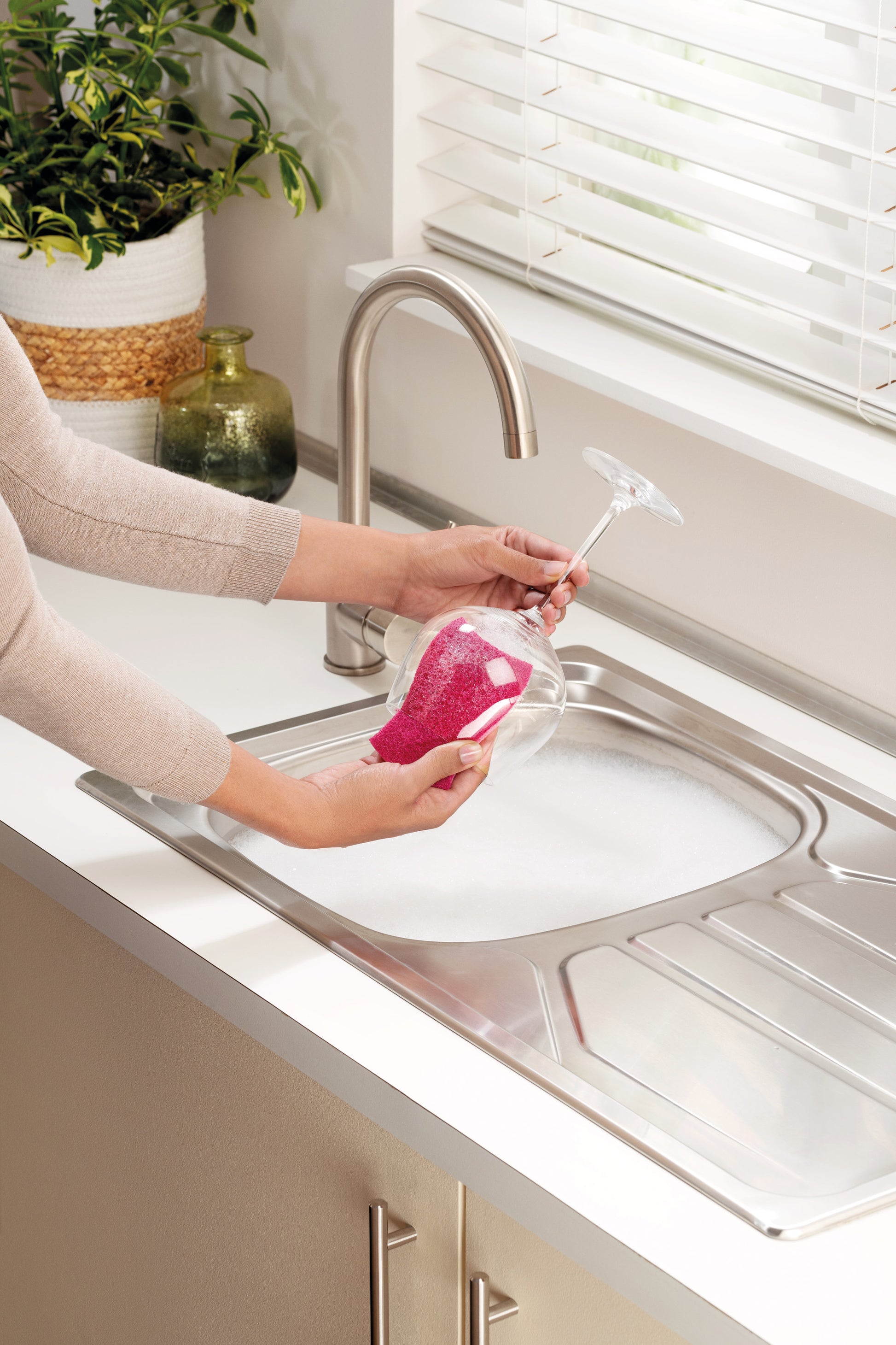 Eine Person wäscht mit einem rosafarbenen Scotch-Brite® Delicate Color kratzfreien Reinigungsschwamm der 3M Deutschland GmbH ein Weinglas unter einem Küchenwasserhahn, während sich im Spülbecken Seifenwasser befindet und auf der Arbeitsplatte eine grüne Topfpflanze steht.