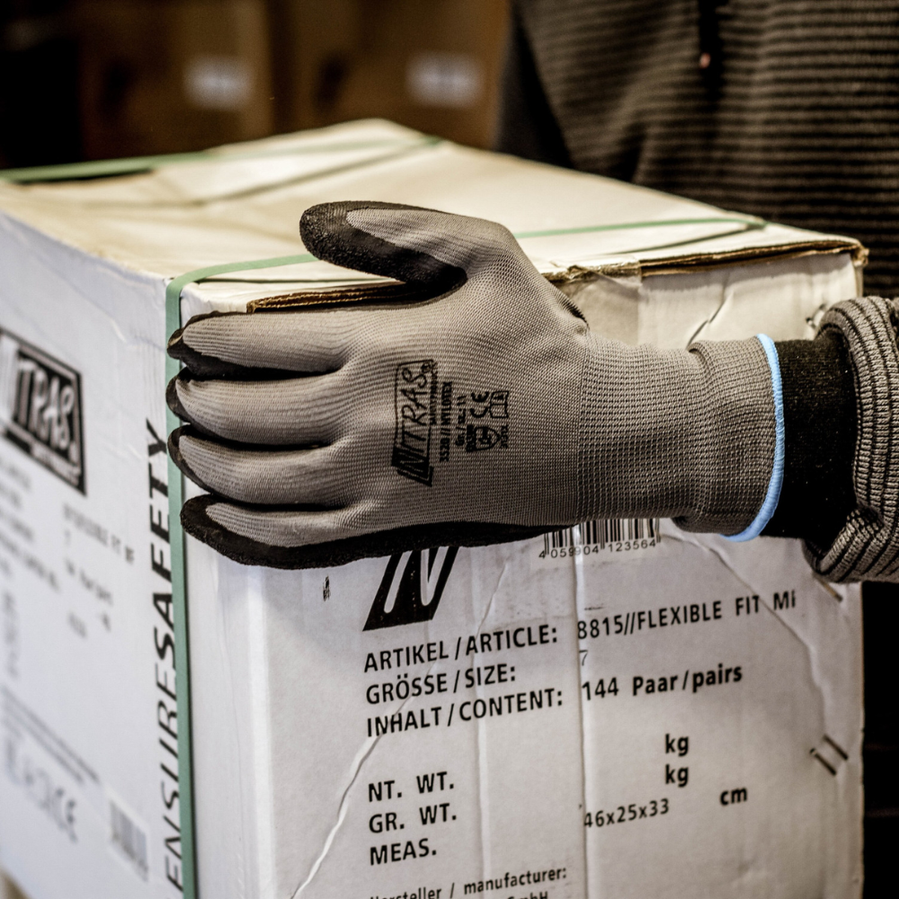 Eine Person, die Nitras Nylotex Nylonhandschuhe mit Latex-Beschichtung der AS Arbeitsschutz GmbH trägt, hält eine große weiße Schachtel mit deutscher und englischer Beschriftung in der Hand; nur ihr Oberkörper und ihr Arm sind teilweise sichtbar.