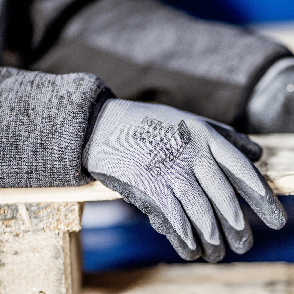 Eine Nahaufnahme zeigt eine Person, die einen strukturierten, grauen Nitras Nylotex Nylonhandschuh mit Latex-Beschichtung der AS Arbeitsschutz GmbH trägt und ihre Hand auf einem Holzbrett abstützt. Auf der Rückseite sind Markierungen und Text zu sehen; der Hintergrund ist leicht unscharf.