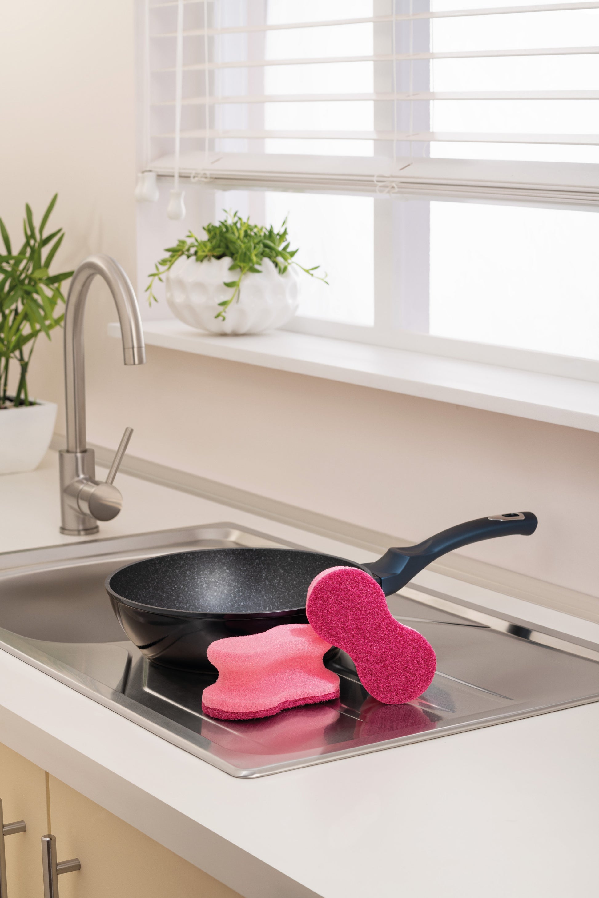 Eine schwarze Bratpfanne liegt auf einer Küchenspüle neben zwei rosafarbenen Scotch-Brite® Delicate Color Comfort kratzfreien Schwämmen (2 pro Packung) der 3M Deutschland GmbH. Im Hintergrund befinden sich Topfpflanzen und ein Fenster mit weißen Jalousien.