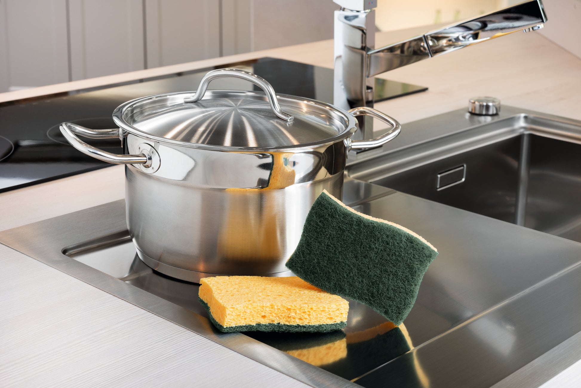 Ein Edelstahltopf mit Deckel steht auf einer Küchenarbeitsplatte neben einer Spüle. Vor dem Topf liegen zwei Scotch-Brite® Classic Reinigungsschwämme der 3M Deutschland GmbH, die auf der Arbeitsplatte liegen.