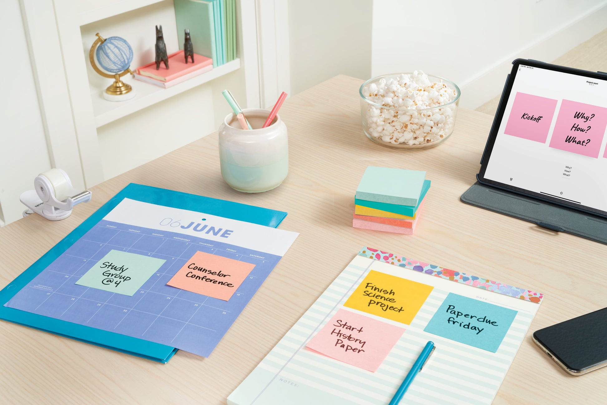 Ein Schreibtisch mit Post-it® Notes Beachside Collection von 3M, einem Juni-Kalender, Erinnerungen, einem Notizbuch, einem Popcornglas, einem iPad mit Notizen, Klebeband, Stiften in einer Tasse und Regalen mit Bürobedarf im Hintergrund.
