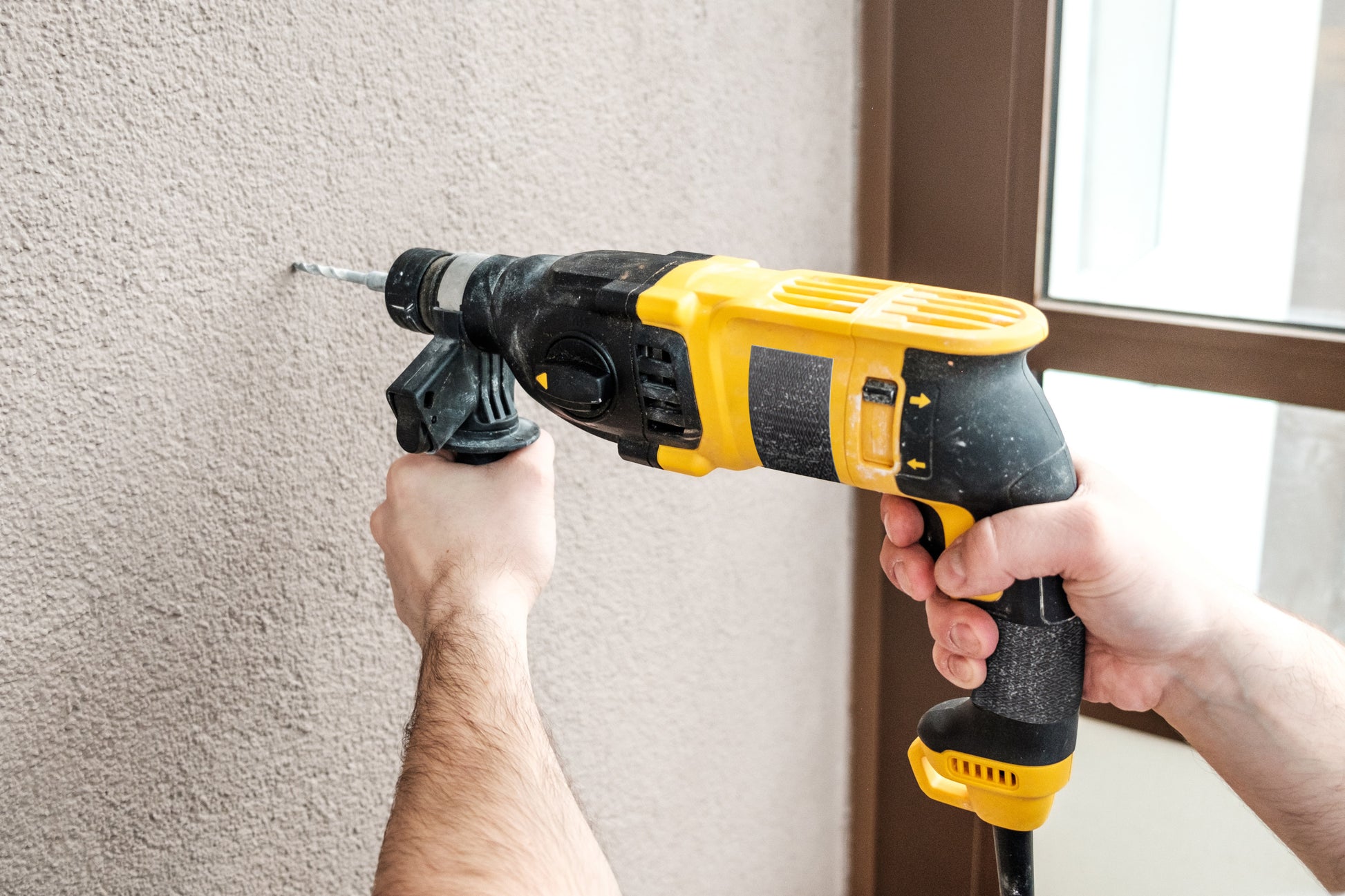 Eine Person bohrt mit einem schwarz-gelben Bohrer in eine beige Wand, während in der Nähe das Scotch® Extremium™ Ultra Hochleistungs-Klebeband DT17 (10 m x 24 mm, 1 Rolle) der 3M Deutschland GmbH zu sehen ist; im Hintergrund sind Hände und ein Teil eines Tür- oder Fensterrahmens zu sehen.