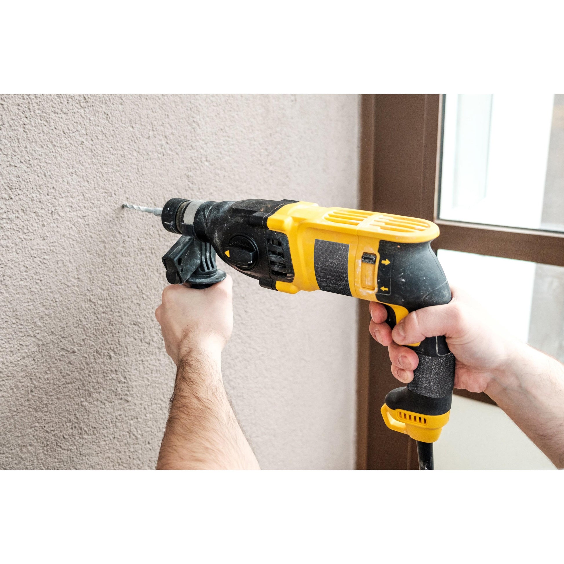 Eine Person bohrt mit einem schwarz-gelben Bohrer in eine beige Wand, während in der Nähe das Scotch® Extremium™ Ultra Hochleistungs-Klebeband DT17 (10 m x 24 mm, 1 Rolle) der 3M Deutschland GmbH zu sehen ist; im Hintergrund sind Hände und ein Teil eines Tür- oder Fensterrahmens zu sehen.