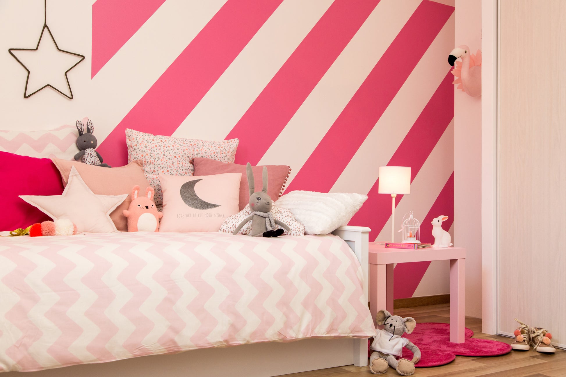 Ein Kinderzimmer mit weißem Bett, rosa und grauen Kissen, Plüschtieren und kräftigen rosa-weiß diagonal gestreiften Wänden, gestrichen mit ScotchBlue™ Malerabdeckband für scharfe Linien von 3M Deutschland GmbH. Eine Lampe, ein Tisch, Spielzeug und Schuhe vervollständigen den gemütlichen Raum.
