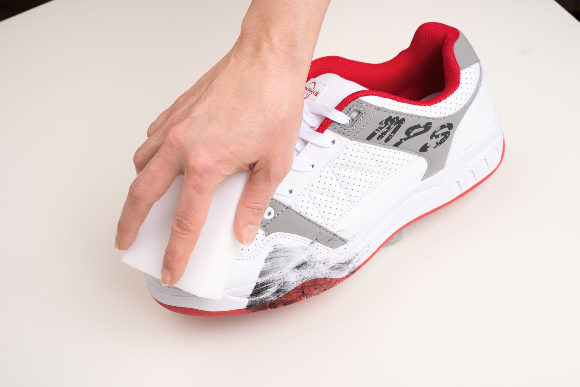 Eine Hand verwendet das Scotch-Brite® Magic Pad von 3M Deutschland GmbH, um schwarze Kratzspuren von der Seite eines weißen Sneakers mit roten und grauen Akzenten auf einer weißen Oberfläche zu entfernen.