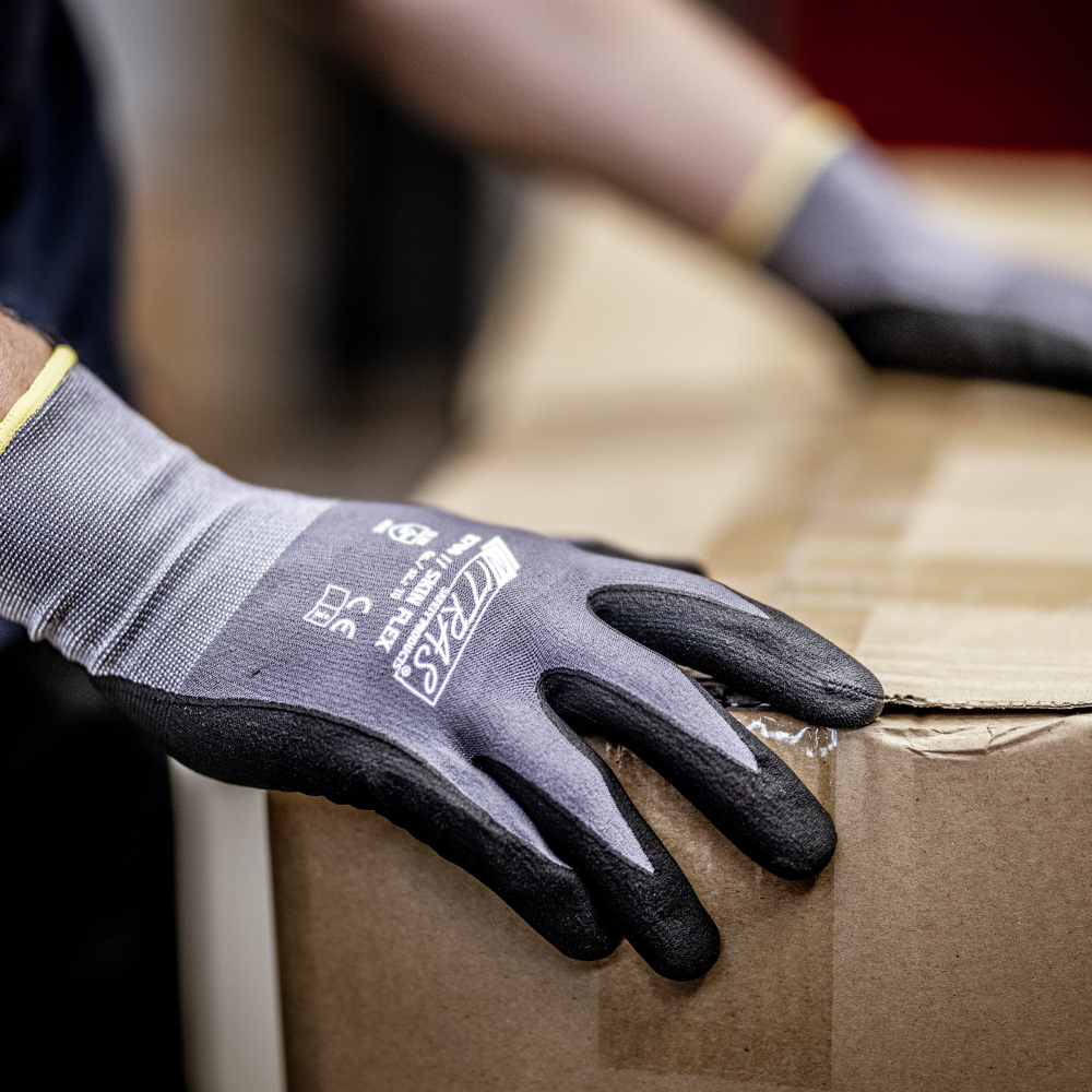 Eine Person trägt NITRAS SKIN FLEX Strickhandschuhe von AS Arbeitsschutz GmbH aus Spezial-Stretch-Strick, grau, teilbeschichtet, hebt einen versiegelten Karton. Sichtbar sind nur die behandschuhten Hände und ein Teil des Kartons.