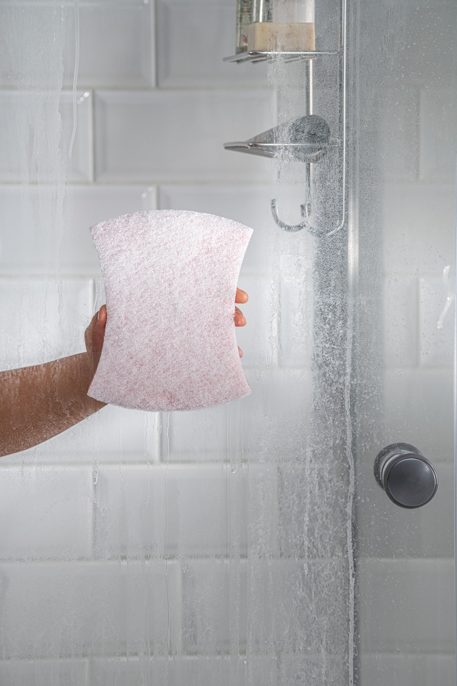 Eine Hand hält den Scotch-Brite® Bath Griffschwamm XXL von 3M Deutschland GmbH in der Dusche, vor nassen weißen Fliesen und einer Glasduschtür mit Wassertropfen. Duschkopf und Seifenablage sind im Hintergrund sichtbar.