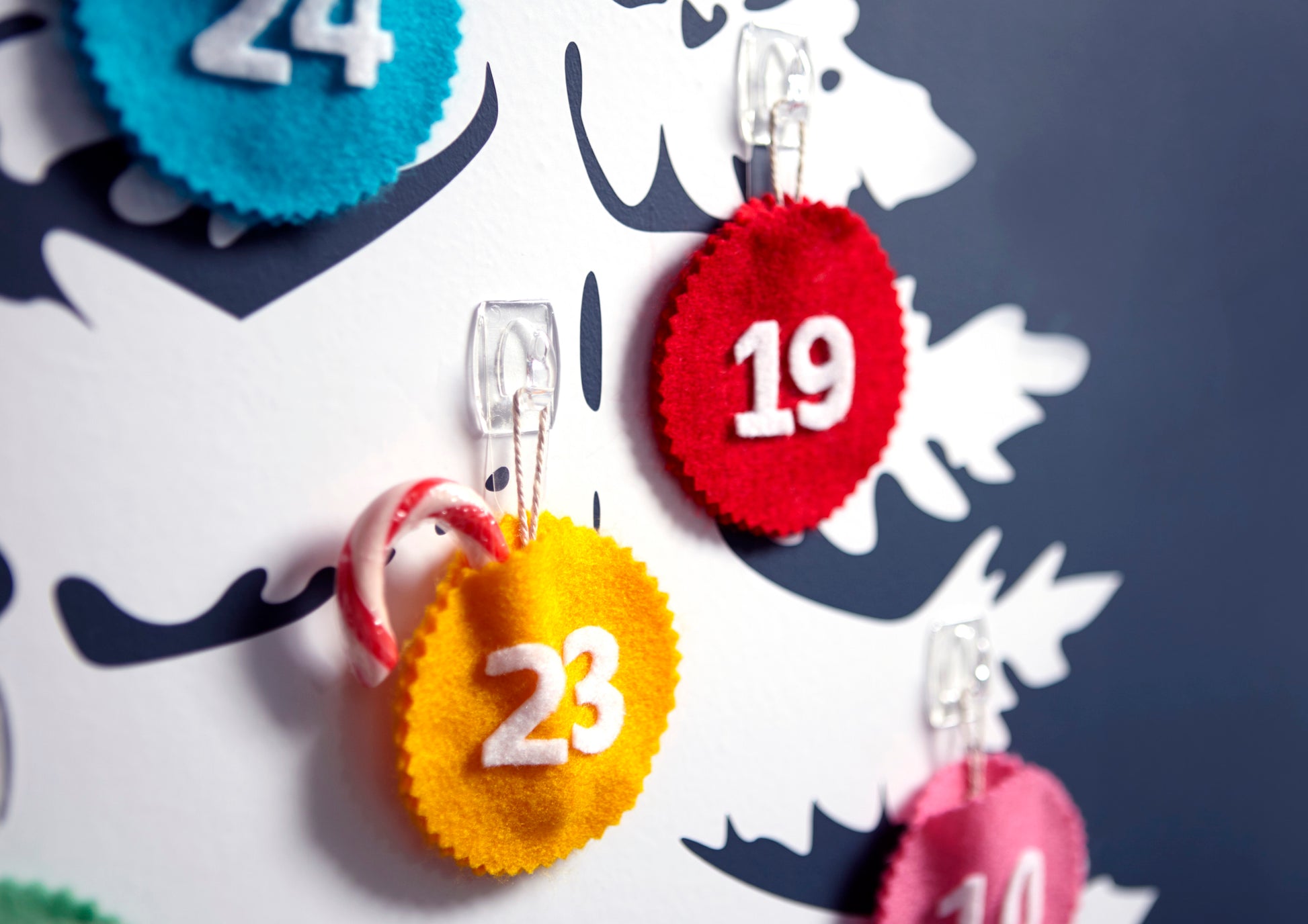 Nahaufnahme einer weißen Tafel mit Filzkreisen mit den Nummern 19, 23 und 24, die jeweils an transparenten Command™ Dekorations-Clips der 3M Deutschland GmbH befestigt sind; an dem Clip der Nummer 23 hängt eine kleine Zuckerstange.